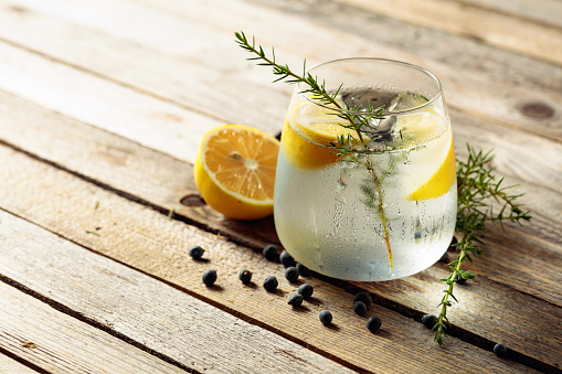 Alcohol drink (gin tonic cocktail) with lemon, juniper branch,  and ice on rustic wooden table, copy space. Iced cocktail drink with lemon and juniper berries.
