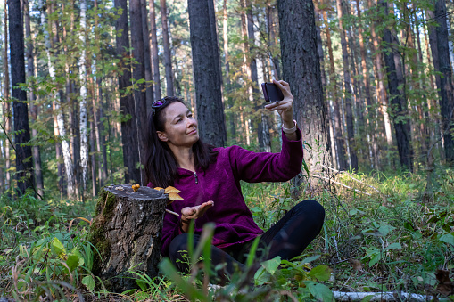 Cheerful woman with honey mushrooms on stump in late summer taking selfie with her smartphone woods in background. Horizontal, copy space