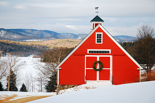 A Christmas wreath hangs on a red barn in a snowy mountain setting in New England