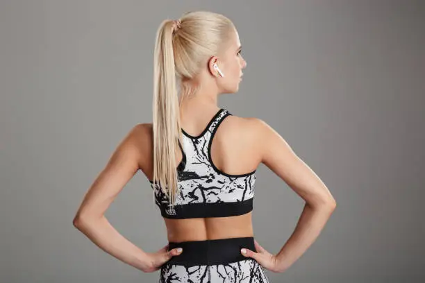 Back view portrait of a blonde young sportsgirl listening to music with earphones while standing isolated over gray background