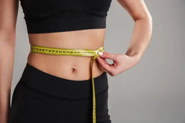 Close up of a slim young sportsgirl holding measuring tape around her waist isolated over gray background