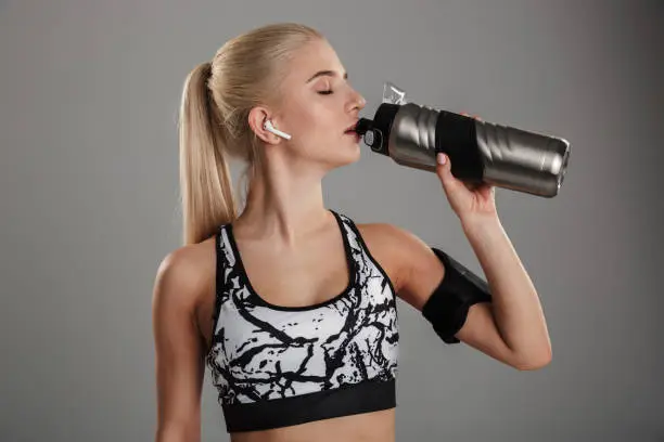 Portrait of a healthy young sportsgirl listening to music with earphones while drinking water isolated over gray background