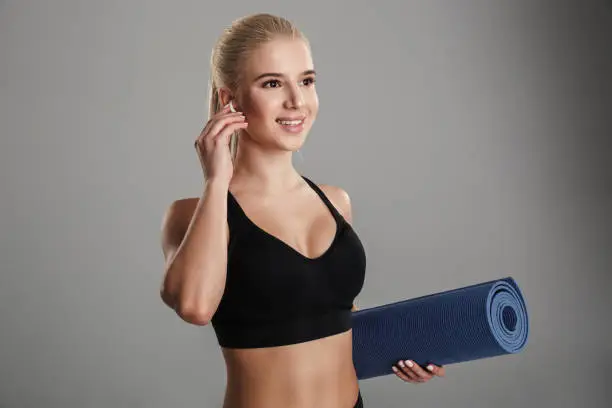 Portrait of a smiling young sportsgirl listening to music with earphones while standing and holding fitness mat isolated over gray background