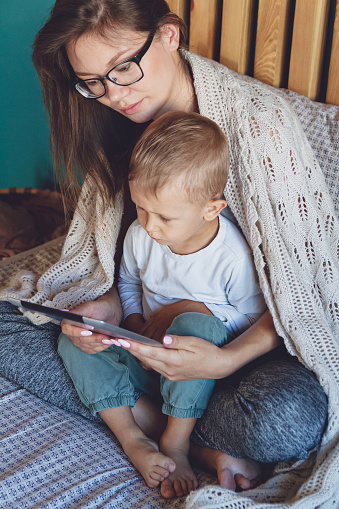 Young mom and son with a tablet under a cozy blanket in the bedroom of their home