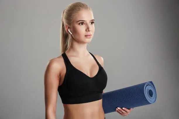 Portrait of a confident young sportsgirl listening to music with earphones while standing and holding fitness mat isolated over gray background