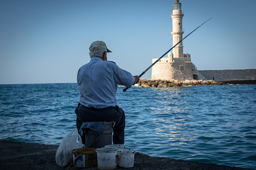 An aged gentleman captured while fishing in the old Venetian port in the city of Chania, Crete. The famous lighthouse is visible in the background.