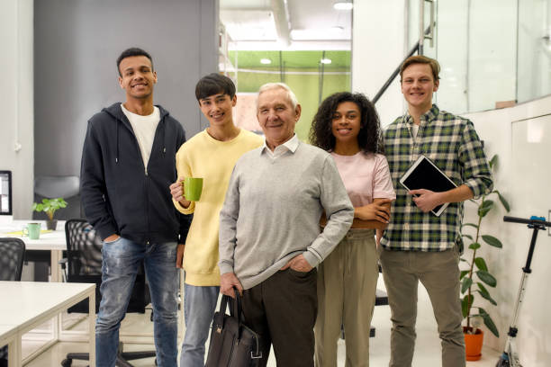 istock Happy diverse team of employees looking at camera while posing with senior intern, standing together in the office, Age diversity in the workplace 1276608608