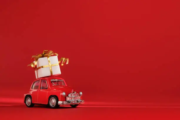 Photo of Red small retro toy car with gift on the roof on red background. Delivery. New Year, Christmas, Valentines Day, World Womans Day, Sale concept