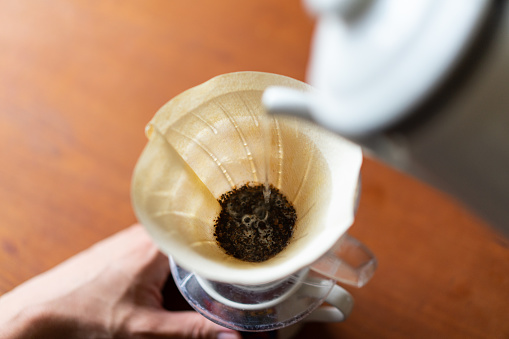 Brewing Coffee at Home - Dripping coffee.\nColorless paper coffee filter.\nEnameled drip kettle (drip pot).\nCone-shaped dripper.\nTaken in natural light. Wooden table.