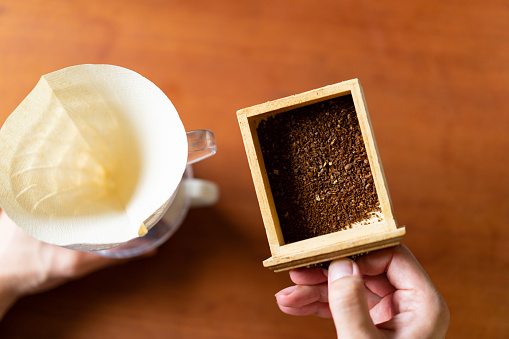 Brewing Coffee at Home - Transfer the coffee powder to the coffee dripper.
Colorless paper coffee filter. Cone-shaped dripper.
Taken in natural light. Wooden table.