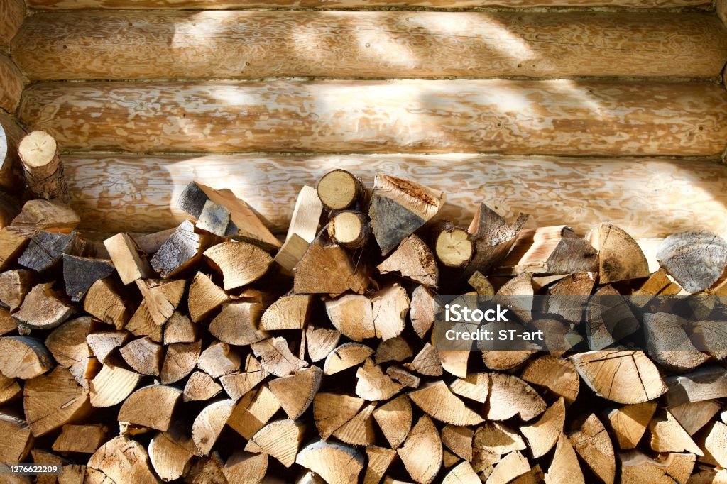 Stacked firewood near the wall of a log village house close-up Stacked firewood near the wall of a log village house close-up. Firewood storage close up. Stocks of wooden logs close-up. Chopped wood and sun glare. Logging in the village. Woodpile with firewood in the village. Countryside lifestyle. Firewood Stock Photo