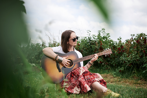 Young brunette hippie woman, wearing boho style clothes, sitting on green grass, holding guitar. Indie musician relaxing on green currant field on sunny summer day. Eco tourism concept.