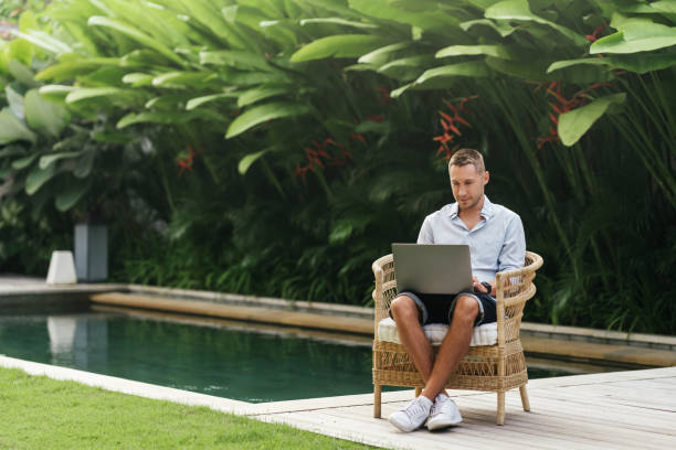 istock Young man using a laptop computer in a garden with a swimming pool. 1276629687