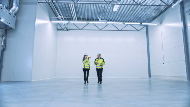 Two Engineers Talking, Walking Across Empty Factory Floor, inspecting, Planning Where All the Machinery Would go. Professionals Industrial Designers Strategizing about New Manufacturing Plant Design