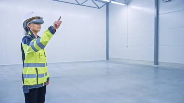 Industry 4.0 Modern Factory: Female Engineer in Empty Warehouse Wearing Augmented Reality Headset Uses Mixed Reality Software and Gestures. Great Mock-up Template for VFX Special Effects and Graphics.