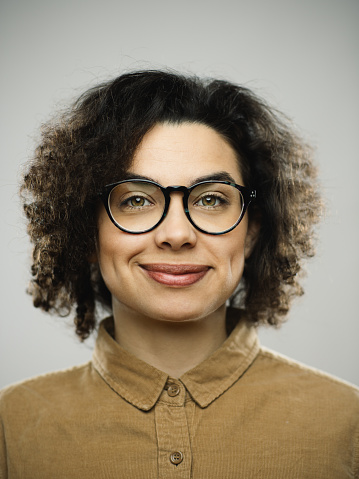 Close-up portrait of young caucasian woman with happy expression against white gray background. Vertical shot of hispanic real people smiling with long curly hair and green eyes. Photography from a DSLR camera. Sharp focus on eyes.