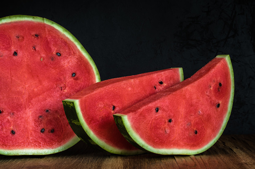 half of a cut ripe watermelon with two large slices and red juicy pulp on an old wooden table on a dark background of black plaster. side view. minimalistic dark still life with copy space