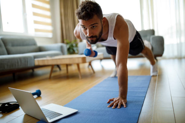 jeune homme utilisant l’ordinateur portatif et exerçant à la maison - home fitness photos et images de collection