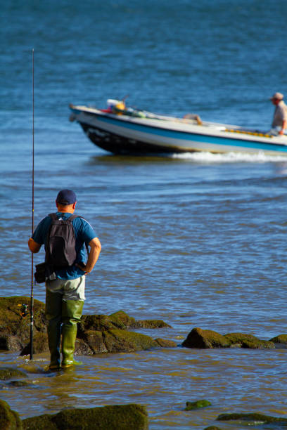 a fisherman wearing long fishing boots , a backpack and a hat is standing on the rock with a long fishing rod - recreational boat nautical vessel fishing rod motorboat imagens e fotografias de stock