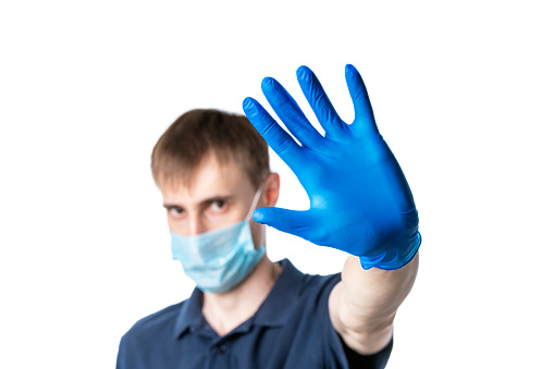A dark-haired man in blue medical gloves and face mask, dressed in a blue t-shirt, shows a stop gesture with one hand isolated on a white background. Concept of protection against Coronavirus