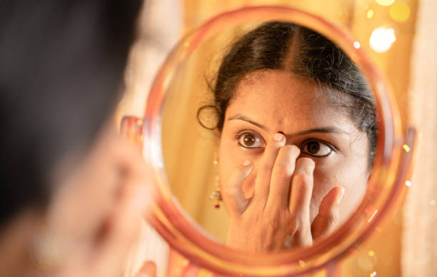 Indian married Woman applying Bindi, sindoor or decorative mark to forehead in front of mirror during festival celebrations with decoration lights as background. Indian married Woman applying Bindi, sindoor or decorative mark to forehead in front of mirror during festival celebrations with decoration lights as background hairstyle bride jewelry women stock pictures, royalty-free photos & images