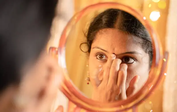 Indian married Woman applying Bindi, sindoor or decorative mark to forehead in front of mirror during festival celebrations with decoration lights as background