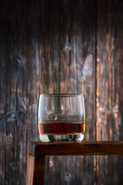 Round glass with a portion of whiskey on the counter against the old wooden wall.