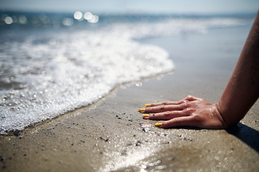 Closeup of the hand of the teenage girl or young woman on the sandy beach. The waves are washing over her hand.\nNikon D850