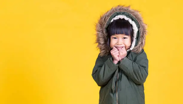 Photo of Portrait of young happy little asian girl wearing fur hooded winter coat sweater isolated on yellow background with copy space. Education childhood lifestyle, winter cold banner