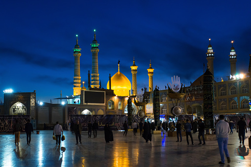 Qom, Iran, 11.08.2016: The Shrine of Fatima Masumeh in the evening with illumination of domes and minarets. People in traditional clothes in the square. Traditional Islamic cityscape.
