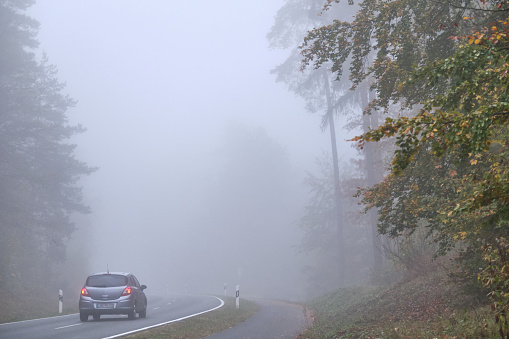 Behringersdorf, Germany - October 24, 2019: A car is driving on a lonesome countryside road with a bicycle path along leading through a forest with dense fog. Seen in October in Germany in Franconia / Bavaria.