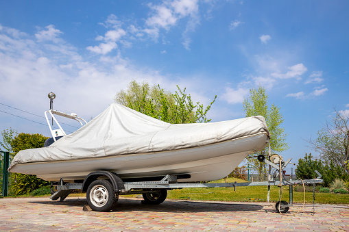 Big modern inflatable motorboat ship covered with grey or white protection tarp standing on steel semi trailer at home backyard on bright sunny day with blue sky on background. Boat vessel storage.