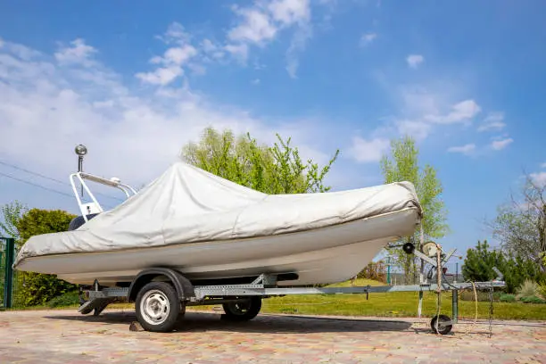Photo of Big modern inflatable motorboat ship covered with grey or white protection tarp standing on steel semi trailer at home backyard on bright sunny day with blue sky on background. Boat vessel storage