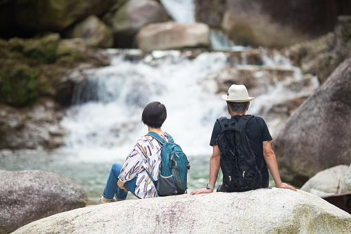 A Japanese senior couple enjoying the outdoors by a waterfall in the mountains of Japan. Sitting on rock.