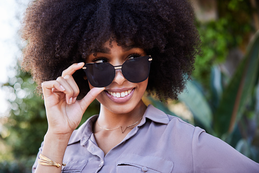 Smiling young African American woman glancing sideways over the rim of her stylish sunglasses while standing outside in the summer