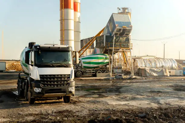 Row many modern big mixer trucks parked against mobile temporary concrete plant factory at new asphalt road construction site early morning day. Heavy machniery and industrial facilities background.