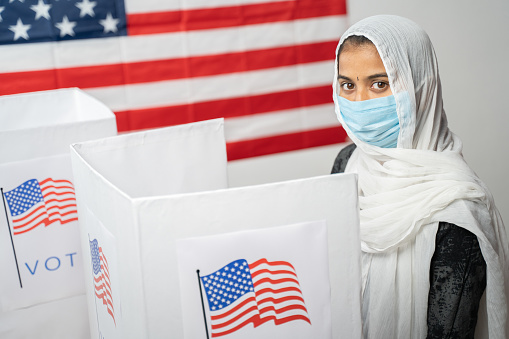 Girl with Hijab or head covering and mask worn at polling booth looking at camera with US flag as background - Concept of US election