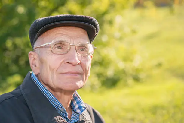 Close up portrait of senior man wearing glasses and cap and sitting on bench in park side view