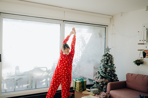 Photo of an excited young woman, jumping for joy in the living room of her apartment; celebrating Christmas in quarantine, and practicing social distancing.