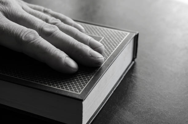 Mature male hand rests on top of a closed book. Hardcover book with gold lettering. Male hand with calluses and cuts. Selective focus.
