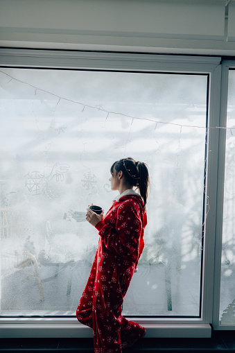 Portrait of a young woman drinking hot chocolate in the living room of her apartment; enjoying the lovely, peaceful Christmas morning; celebrating Christmas in quarantine, and practicing social distancing.