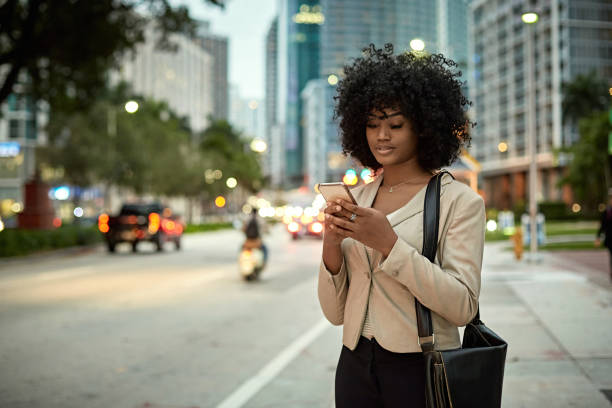 African-American Businesswoman Outdoors in Downtown Miami Young female corporate executive standing at curb of busy street checking smart phone as she waits for crowdsourcing taxi at dusk. crowdsourced taxi photos stock pictures, royalty-free photos & images