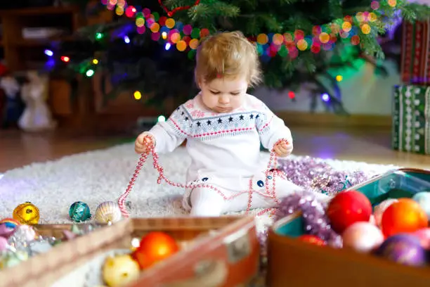 Adorable baby girl holding colorful lights garland in cute hands. Little child in festive clothes decorating Christmas tree with family. First celebration of traditional holiday called Weihnachten.