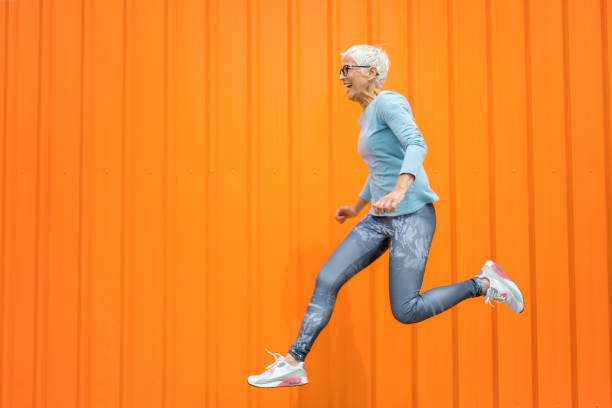 glückliche seniorin springt vor orangefarbenem hintergrund. - hochspringen stock-fotos und bilder