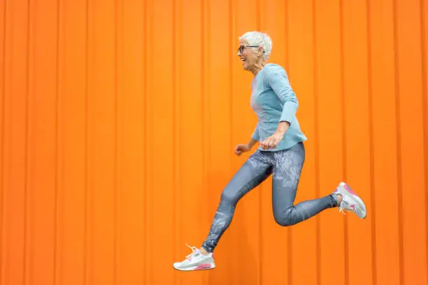 Portrait of beautiful excited mature woman with grey hair jumping . Attractive middle aged woman with beautiful smile isolated over orange background.