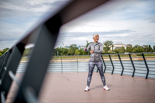 Beautiful athletic mature woman in sportswear stretching her legs after morning jogging by the river.