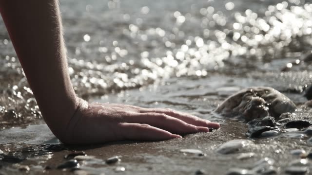The young woman by the sea sits with her hands on the sand and pebbles, and the waves flow over her hand.