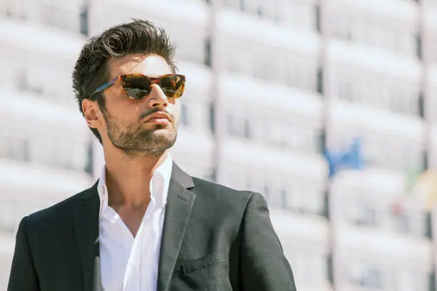 Sexy gorgeous stylish man. Sunglasses. City style. A beautiful and charming man with sunglasses outdoors. Stubble and blacks hair. Intense light. White shirt and stylish jacket.