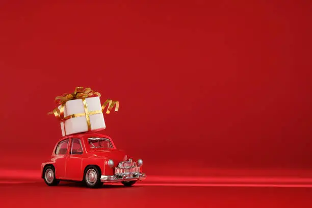 Photo of Red small retro toy car with gift on the roof on red background. Delivery. New Year, Christmas, Valentines Day, World Womans Day, Sale concept