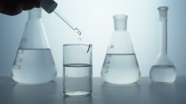 Close-up of a pipette in hand and a glass beaker against the background of flasks in the laboratory.